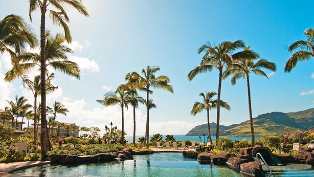 A clear pool with palm trees, a view of the ocean, and a mountainous backdrop.