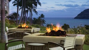 An outdoor seating area with stone tables and chairs, a fire feature, and a view of the ocean and palm trees.