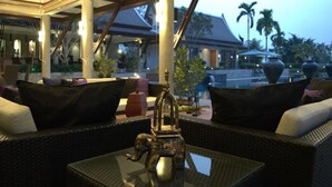 An outdoor seating area with wicker furniture and a glass table, overlooking a pool and palm trees. - DDM Siam Thailand (Bang Lamung)