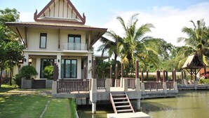 A two-story house with a balcony, a small bridge over a water body, and palm trees in the background. - DDM Siam Thailand (Bang Lamung)