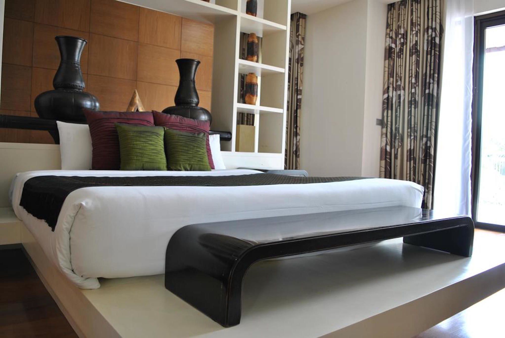 A modern hotel room with a large bed, a wooden headboard, a built-in shelf unit, and a large window with curtains.