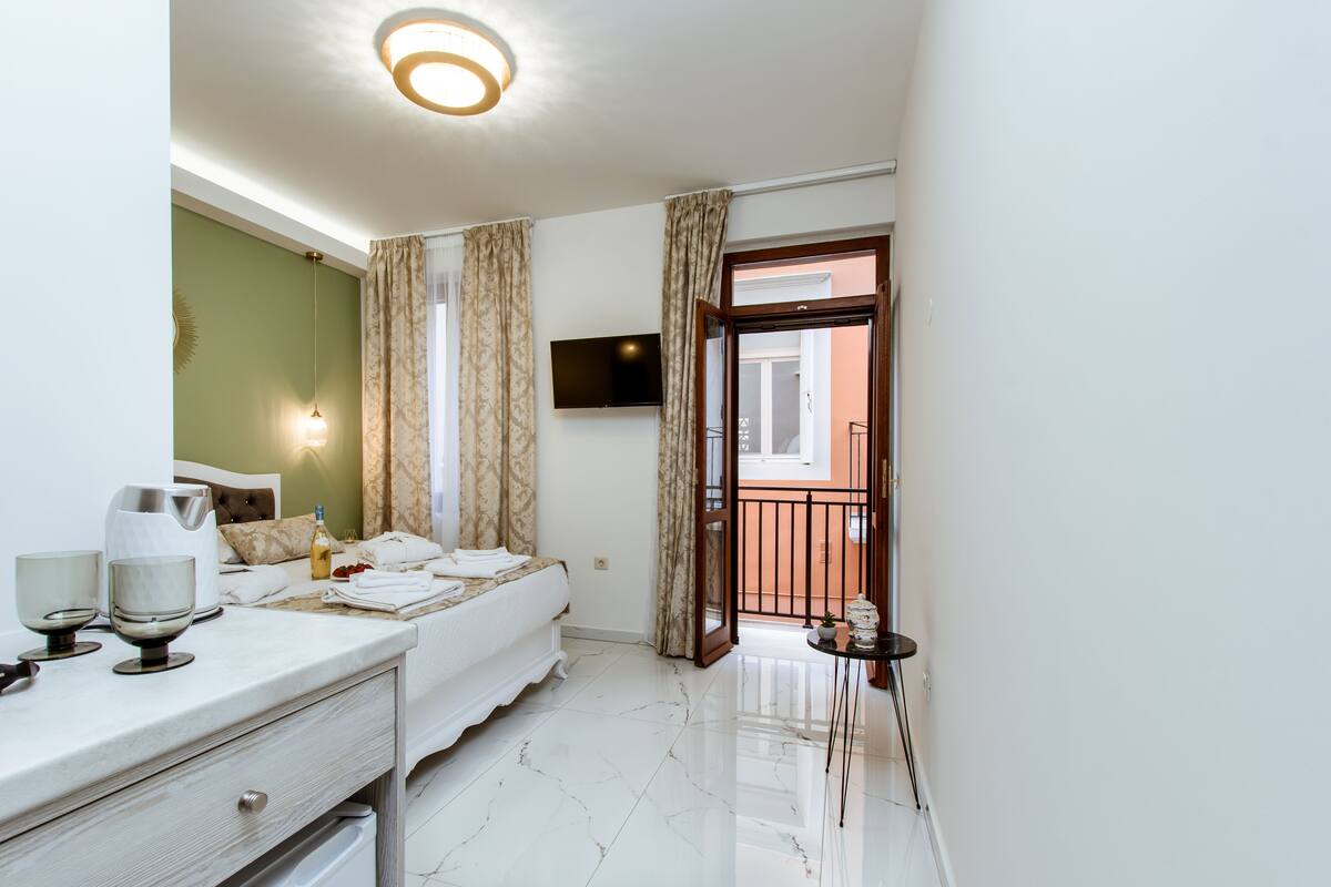 Une chambre d’hôtel avec un lit, une table de chevet avec des verres et une bouteille, un téléviseur fixé au mur et un balcon avec une rambarde.