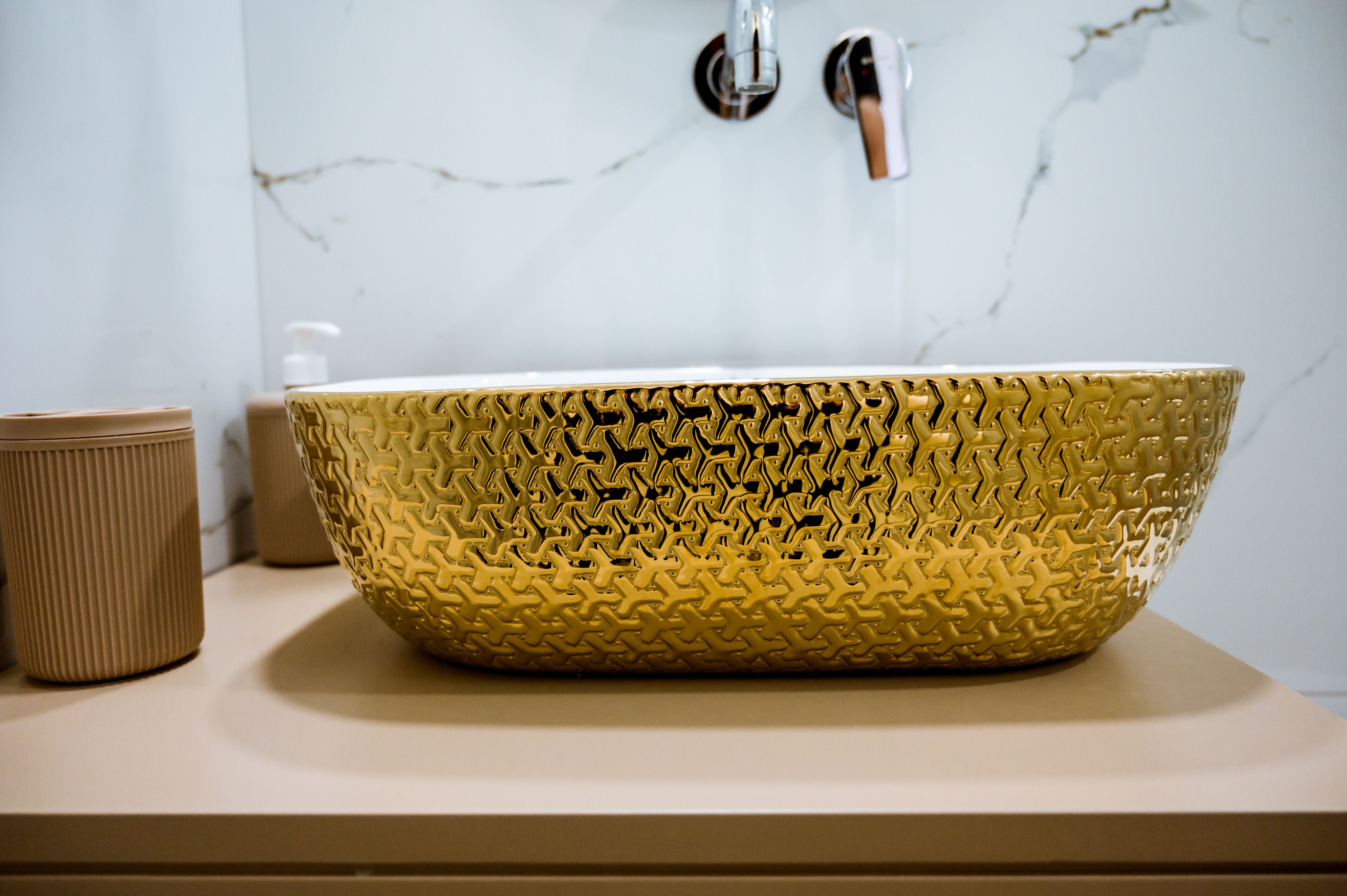 Un baño con un lavamanos texturizado dorado, una encimera color beige y una pared de mármol blanco.