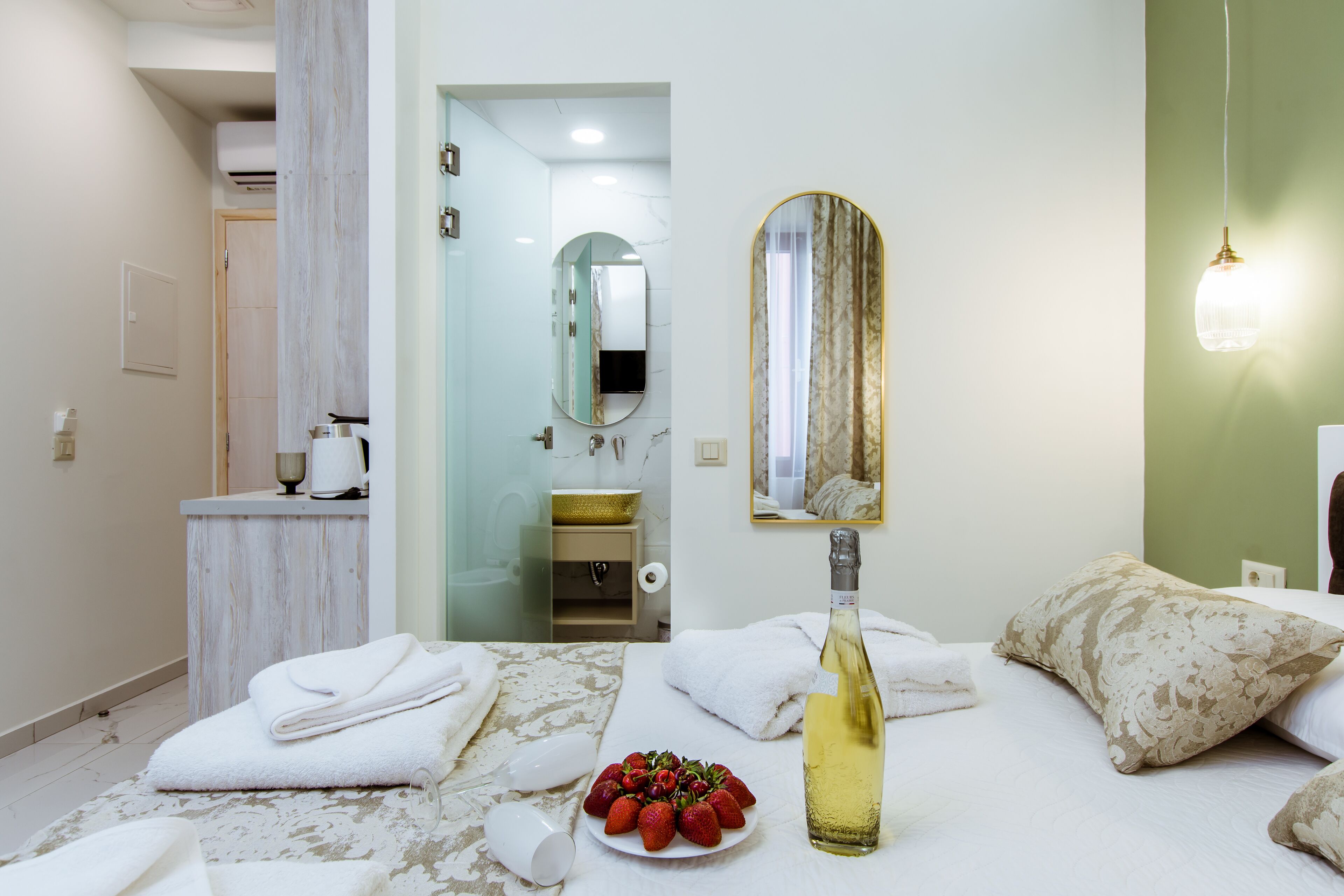 A hotel room with a bed, a bedside table with a bottle and strawberries, a mirror, and a bathroom visible in the background.