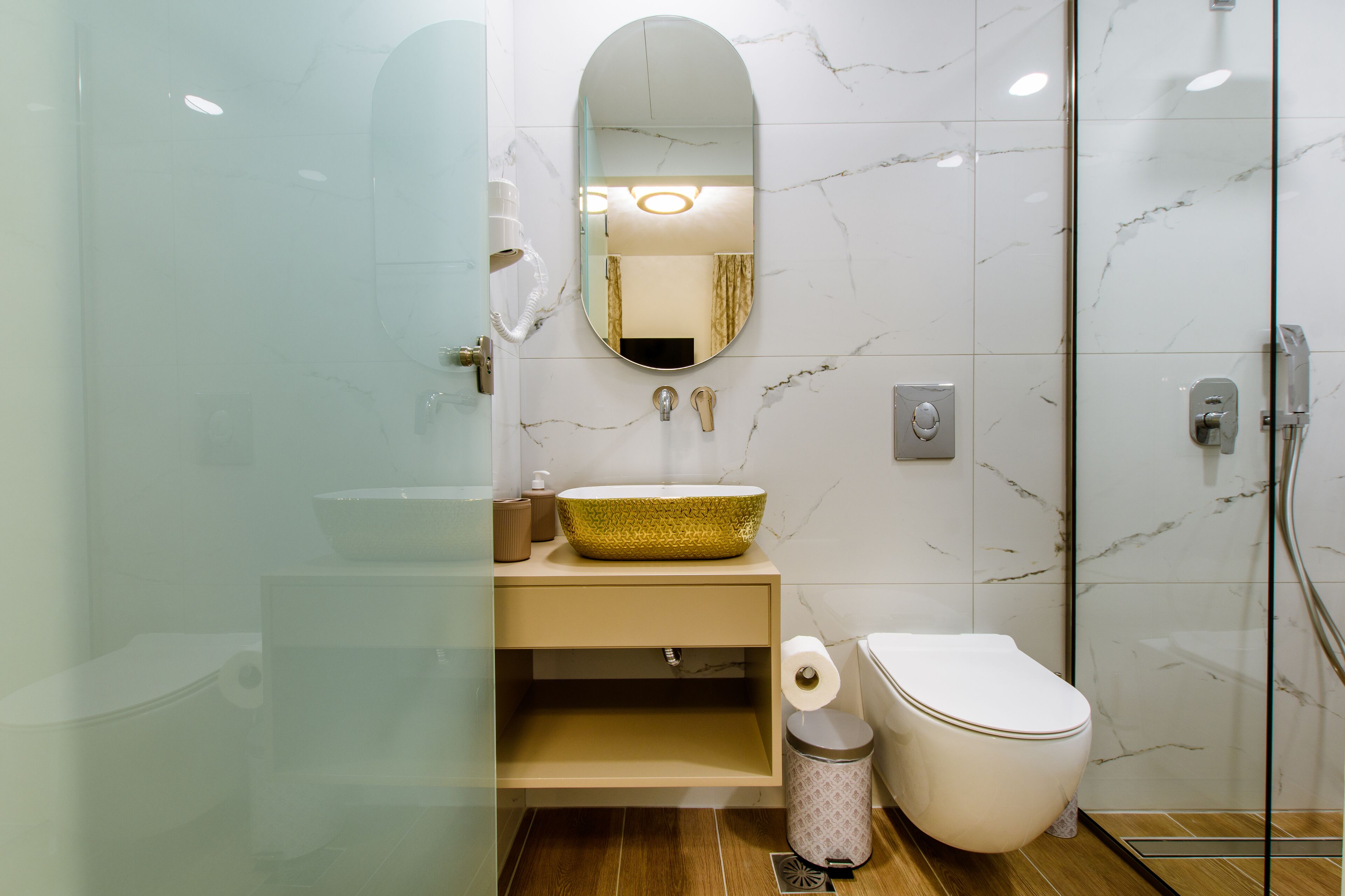 A modern bathroom with a marble backsplash, a glass-enclosed shower, a beige vanity with a gold bowl sink, and a toilet with a paper towel dispenser.