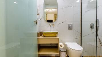 A modern bathroom with a marble backsplash, a glass-enclosed shower, a beige vanity with a gold bowl sink, and a toilet with a paper towel dispenser.