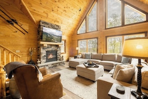 A cozy log cabin interior with a stone fireplace, a large TV mounted on the wall, and a comfortable seating area. - A Lil Slice of Heaven by Taylor Made Deep Creek (McHenry)