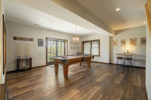 A pool table in a room with wooden flooring, a ceiling light, and a bar area with stools. - A Lil Slice of Heaven by Taylor Made Deep Creek (McHenry)