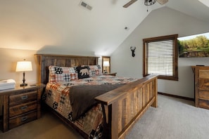 A bedroom with a wooden bed, a nightstand, a lamp, a window with blinds, and a deer head decoration on the wall. - A Lil Slice of Heaven by Taylor Made Deep Creek (McHenry)