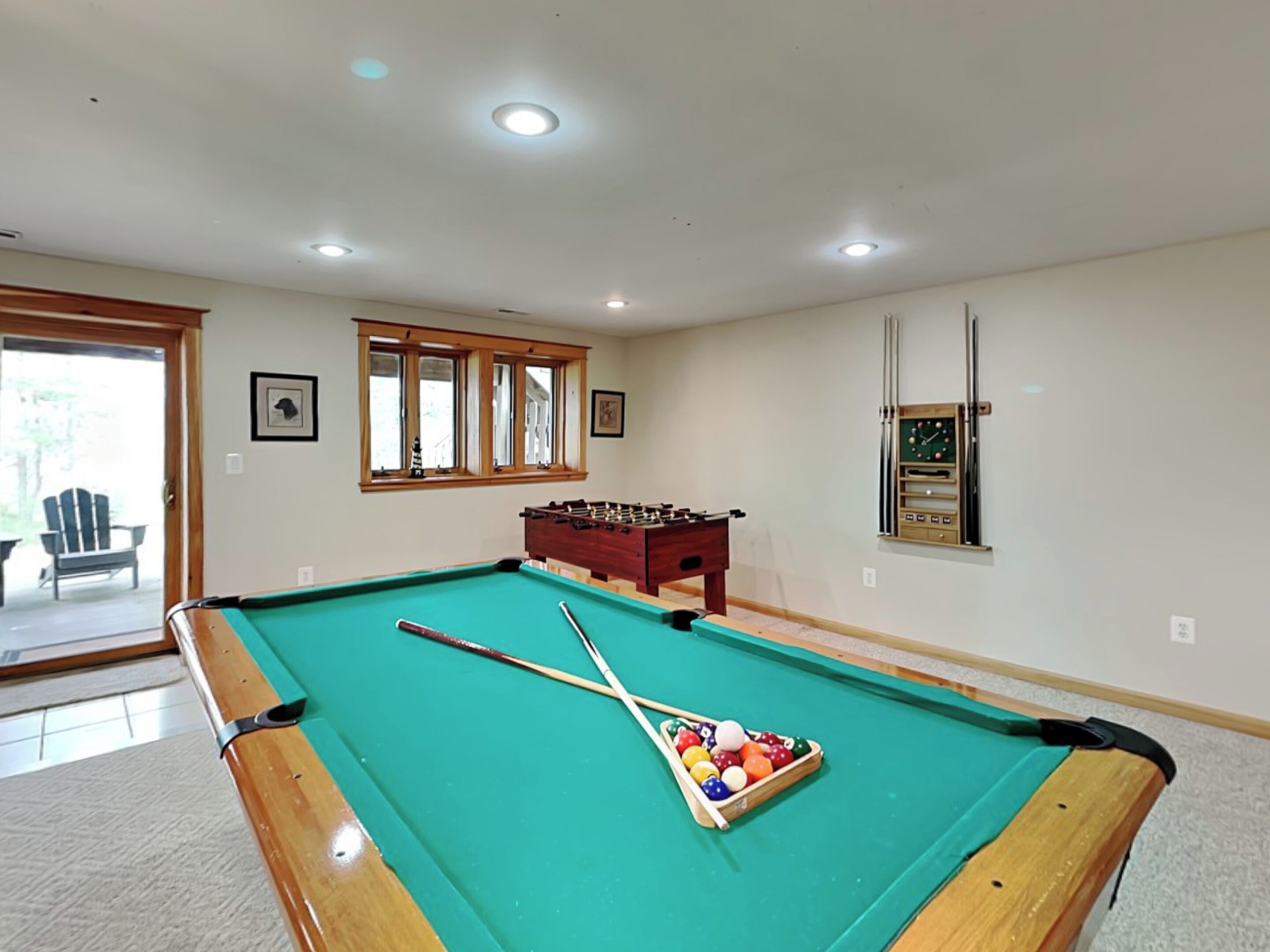 A pool table with pool cues and balls, a foosball table, and a clock on the wall.
