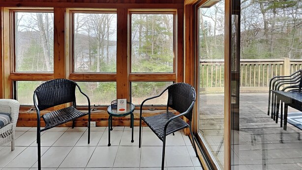 Une terrasse couverte avec deux chaises en métal noir et une petite table, de grandes fenêtres offrant une vue sur les arbres et une terrasse.