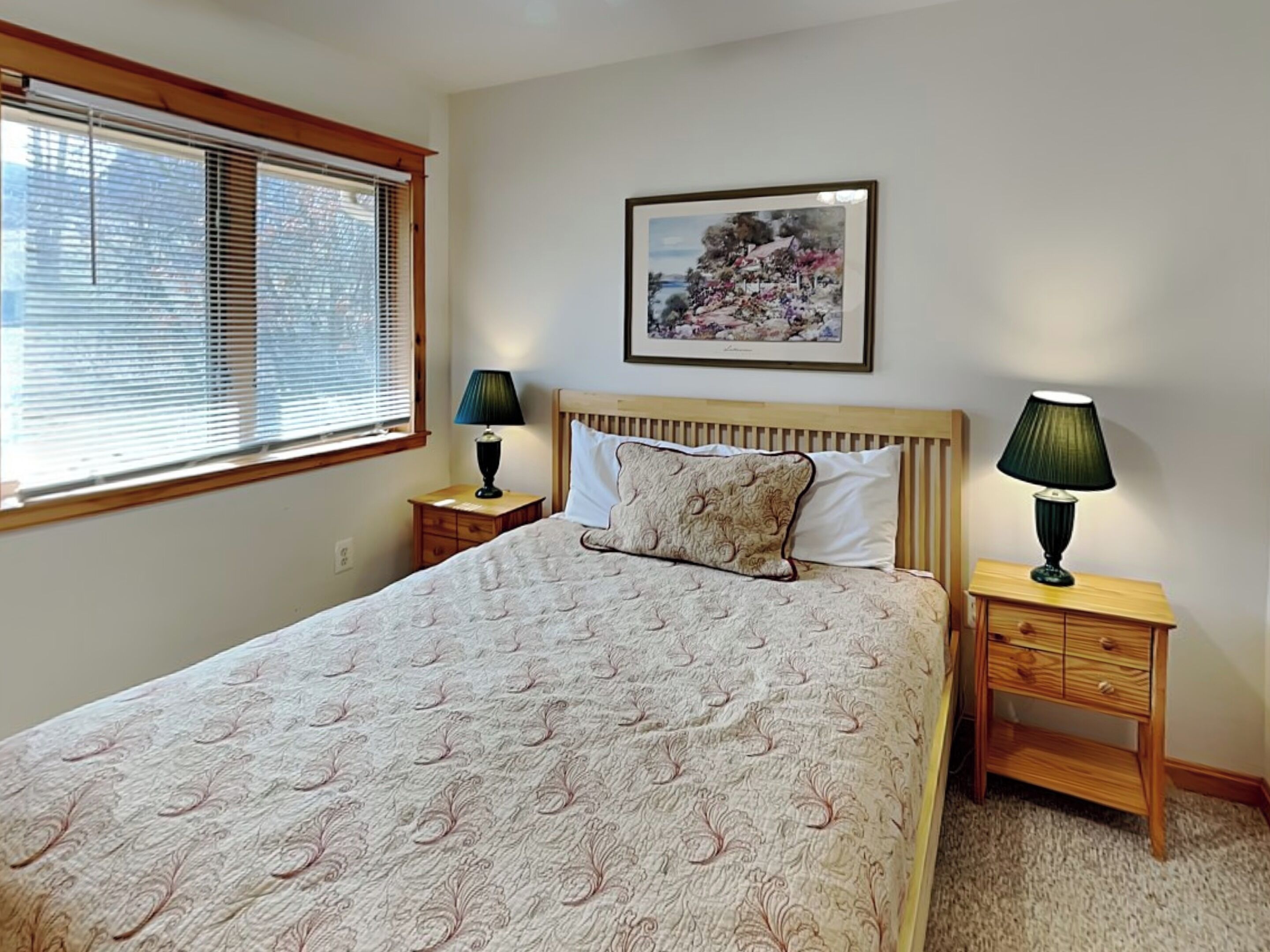 A bedroom with a bed, two bedside tables, a window with blinds, and a framed picture on the wall.