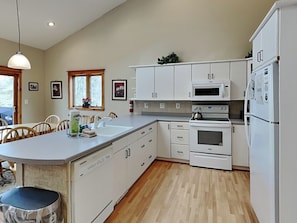 Una cocina moderna con armarios blancos, horno empotrado, microondas y lavavajillas. Cuenta con un comedor con mesa y sillas, y una ventana con vistas a árboles.