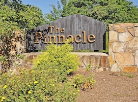 A wooden sign with the text "The Pinnacle" against a stone wall backdrop, surrounded by greenery.