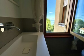 Et badeværelse med et hvidt håndvask, et spejl og havudsigt fra et vindue.