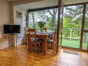 Un coin repas avec une table et des chaises en bois, un téléviseur posé sur un meuble, et une vue sur un jardin verdoyant à travers de grandes fenêtres.