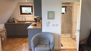 Una cocina compacta con fregadero, tostadora, hervidor y microondas. Un sillón gris está en primer plano. Se ve una zona de ducha a través de una puerta abierta.