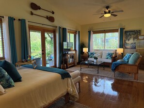 Une pièce spacieuse comprenant un lit, un canapé, une table basse et un téléviseur. Elle est dotée de rideaux, d’un ventilateur de plafond et offre une vue sur la verdure environnante.