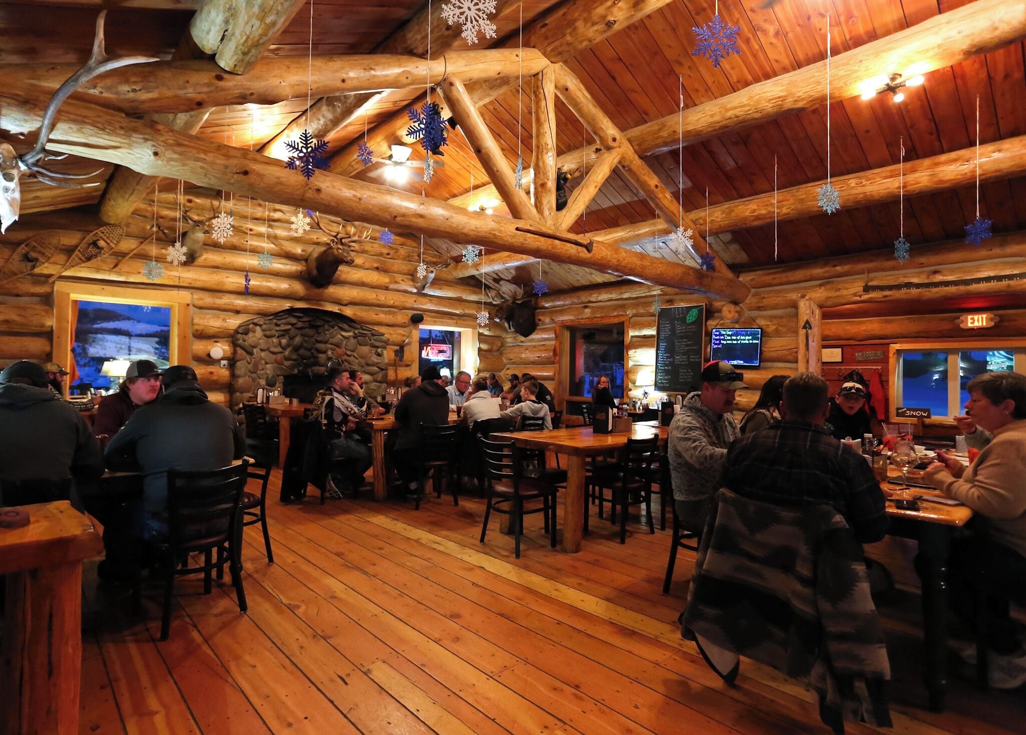 A log cabin-style restaurant with wooden beams and a stone fireplace. Tables are set for dining, and snowflake decorations hang from the ceiling.
