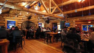 A log cabin-style restaurant with wooden beams and a stone fireplace. Tables are set for dining, and snowflake decorations hang from the ceiling.