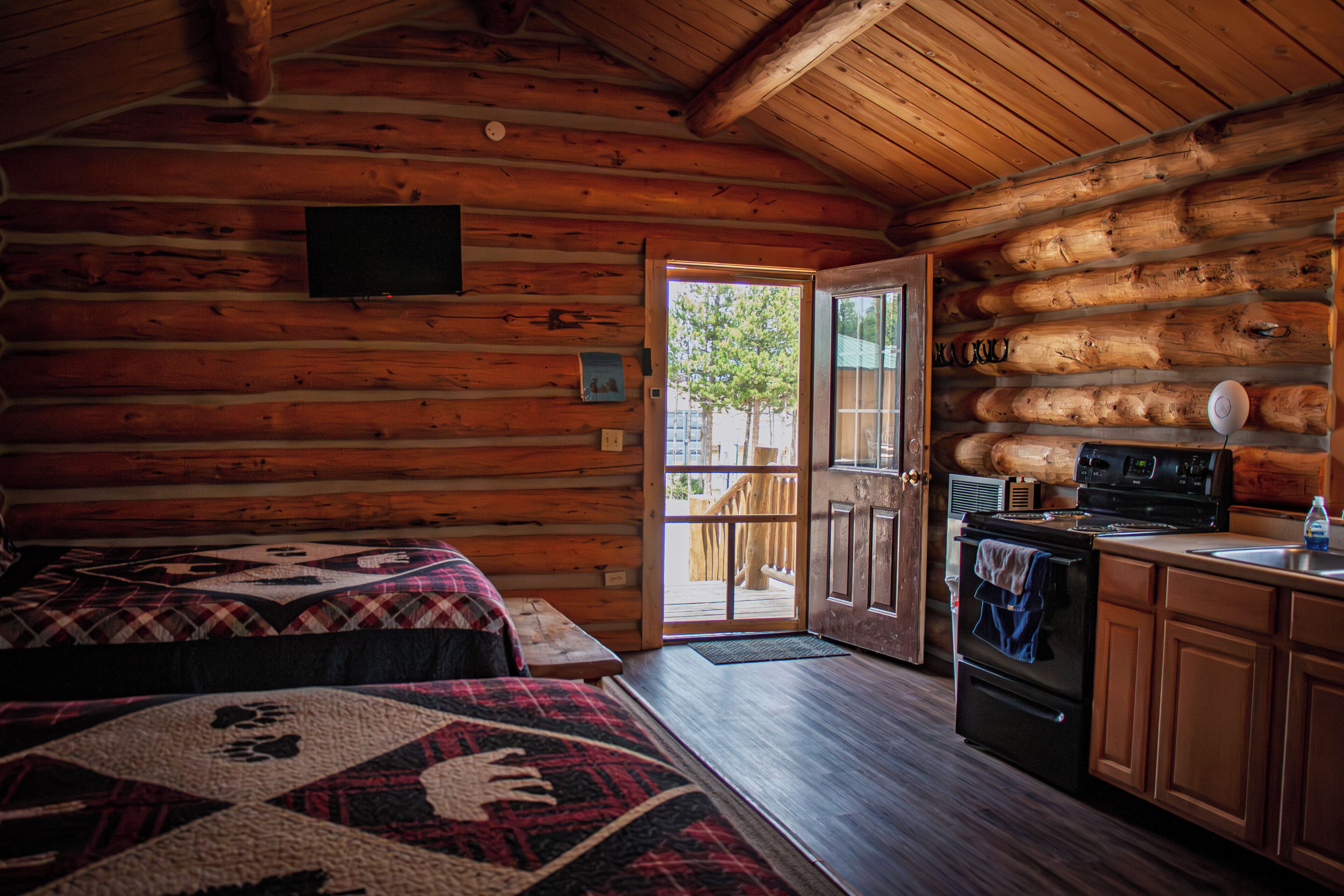 A log cabin interior with two beds, a kitchen, and a sliding glass door leading to a balcony.