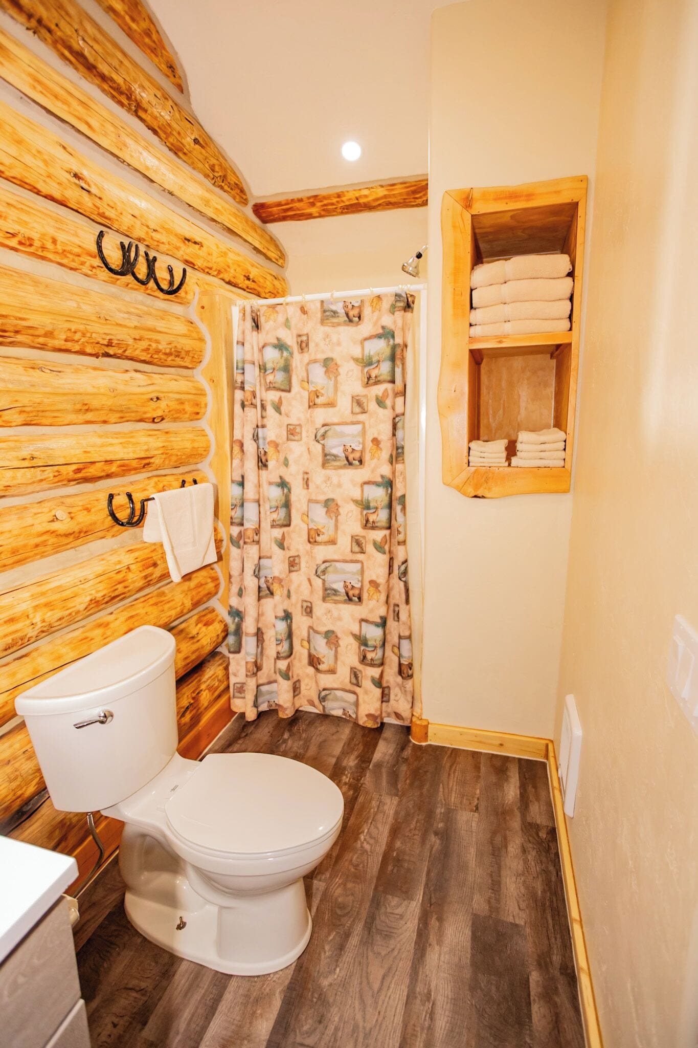 A bathroom with a wooden log wall, a white toilet, a wooden shelf with towels, and a shower curtain with a pattern.