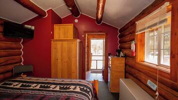 A log cabin bedroom with a bed, a wooden wardrobe, a television, and a window with blinds.