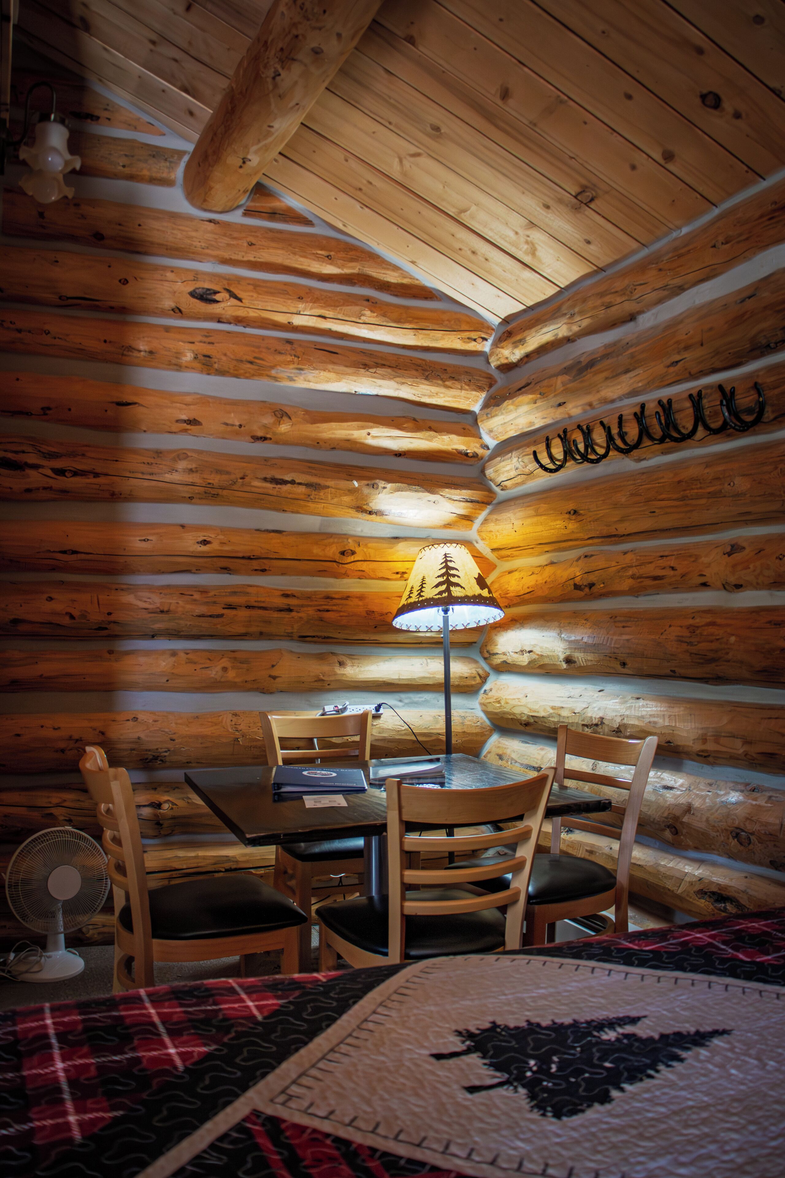 L’intérieur d’un chalet accueillant, avec un mur en rondins de bois, une table avec des chaises, une lampe et un ventilateur.