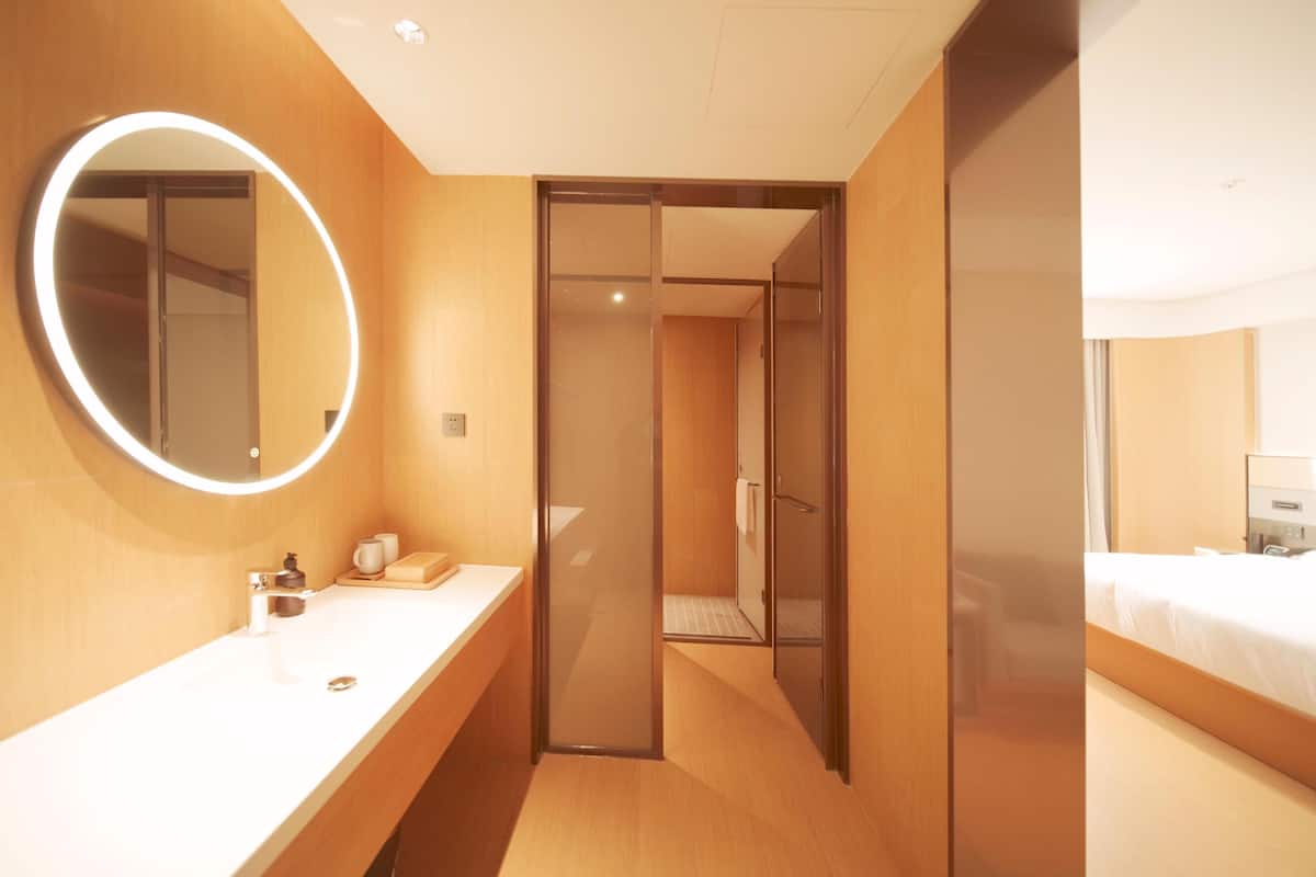 A hotel room with a wooden vanity, a round mirror with a light, a shower area, and a bed.