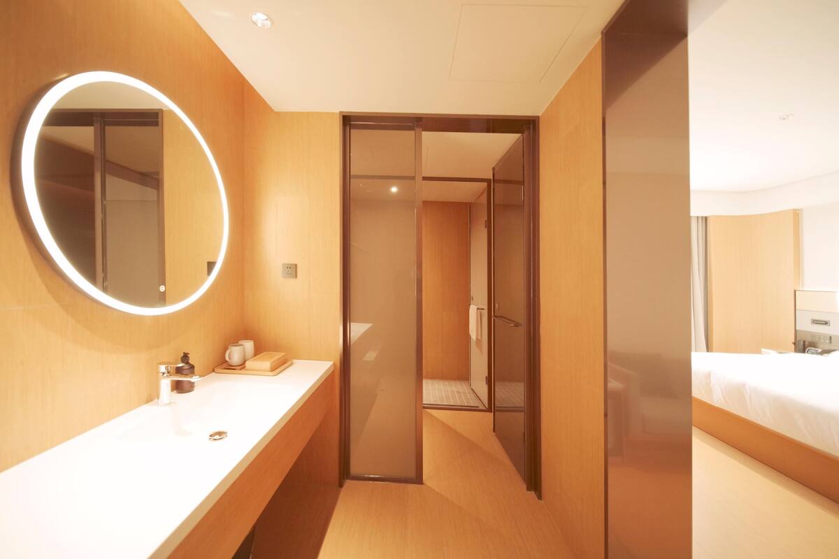 A hotel room with a wooden vanity, a round mirror with a light, a shower area, and a bed.