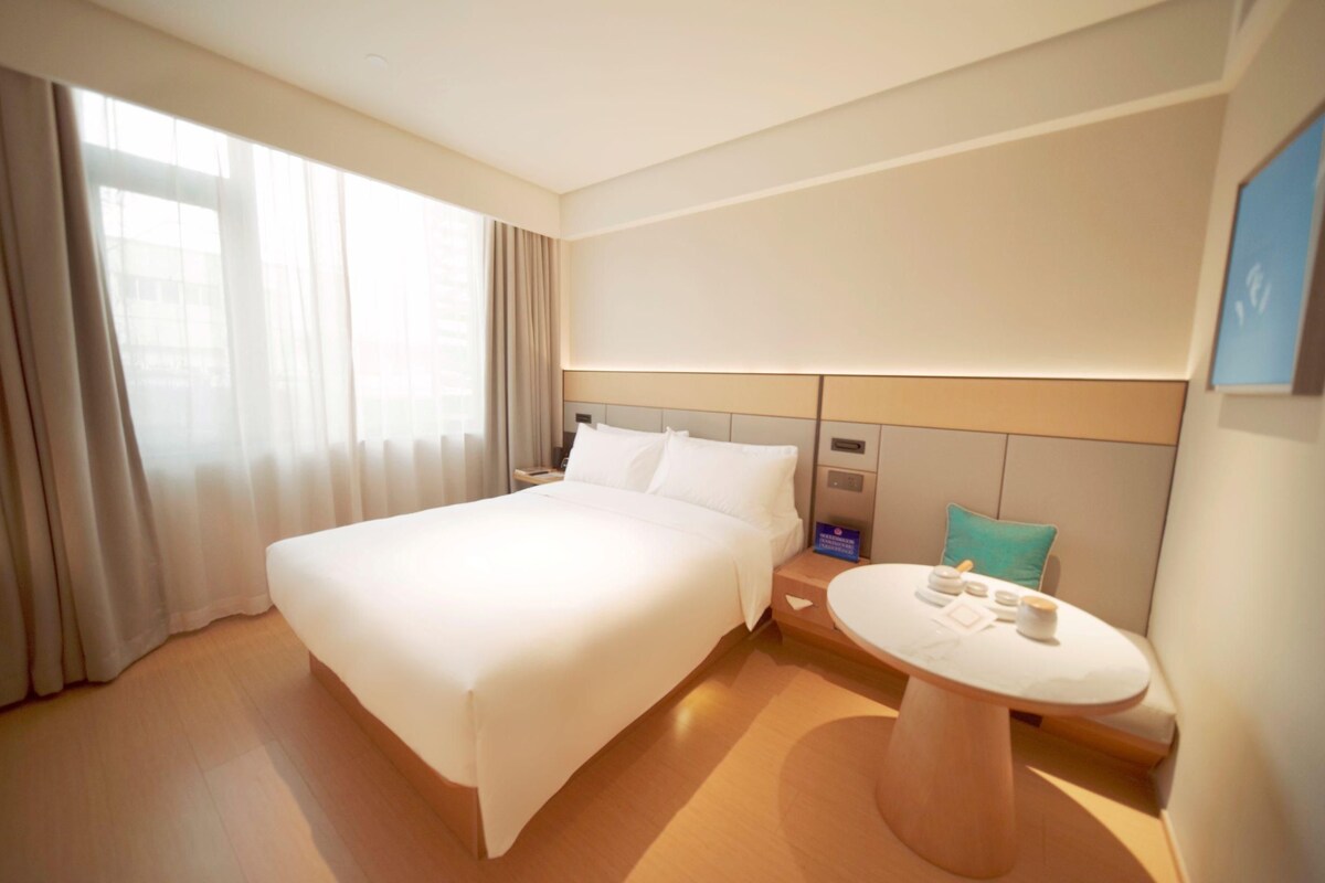 A hotel room with a large bed, a bedside table with a lamp, a round table with a chair, and a view of the city through the window.
