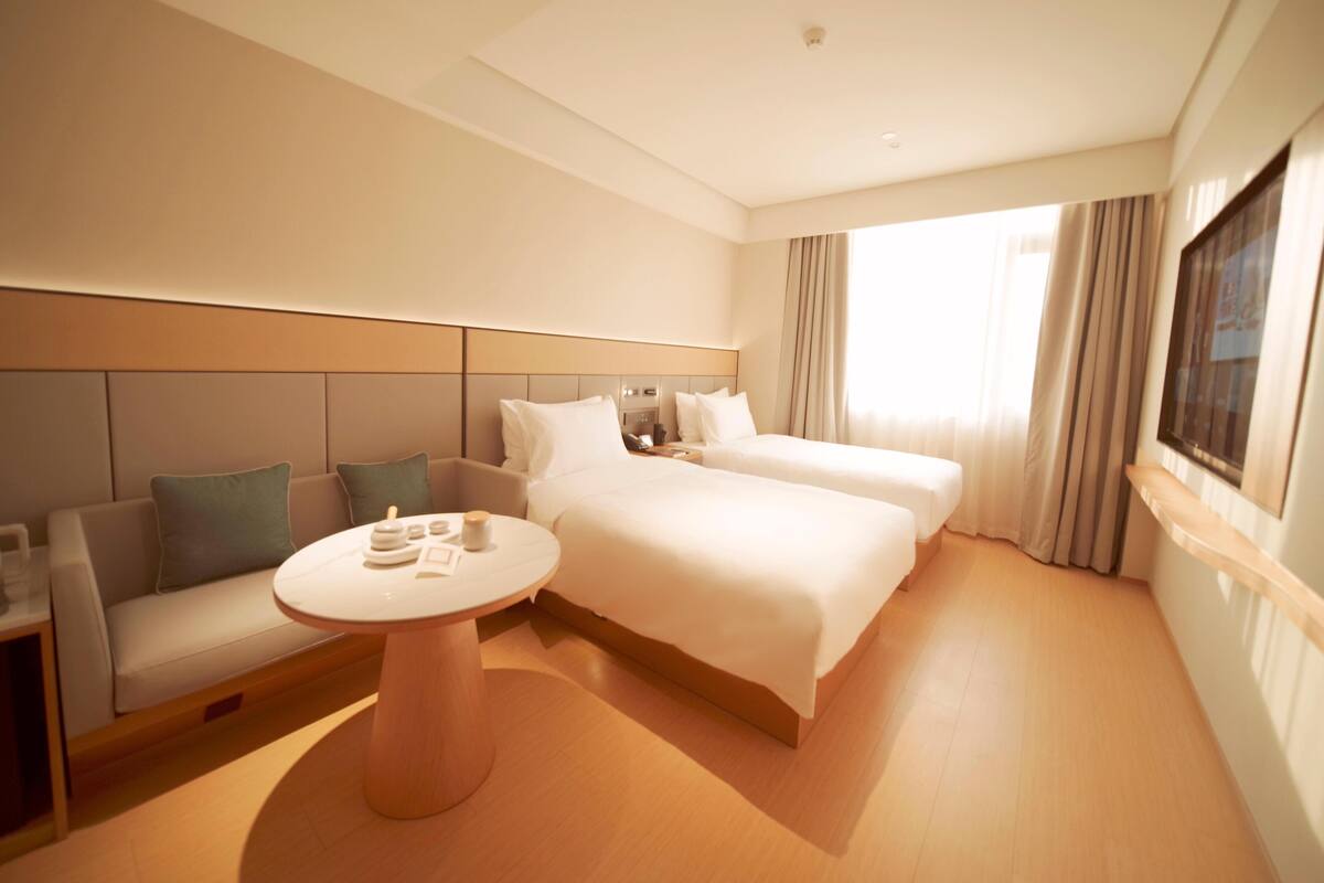 A hotel room with a sofa, two beds, a round table, and a large window with curtains.