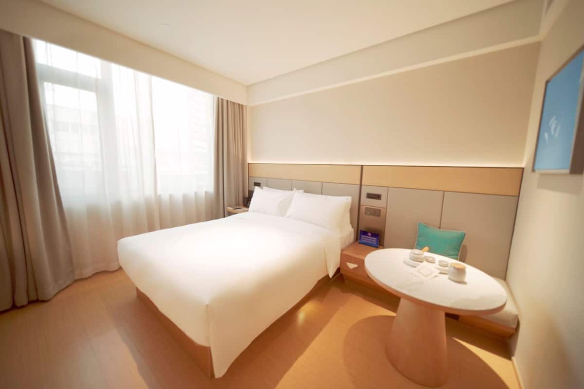 A hotel room with a large bed, a bedside table with a lamp, a round table with a chair, and a view of the city through the window.