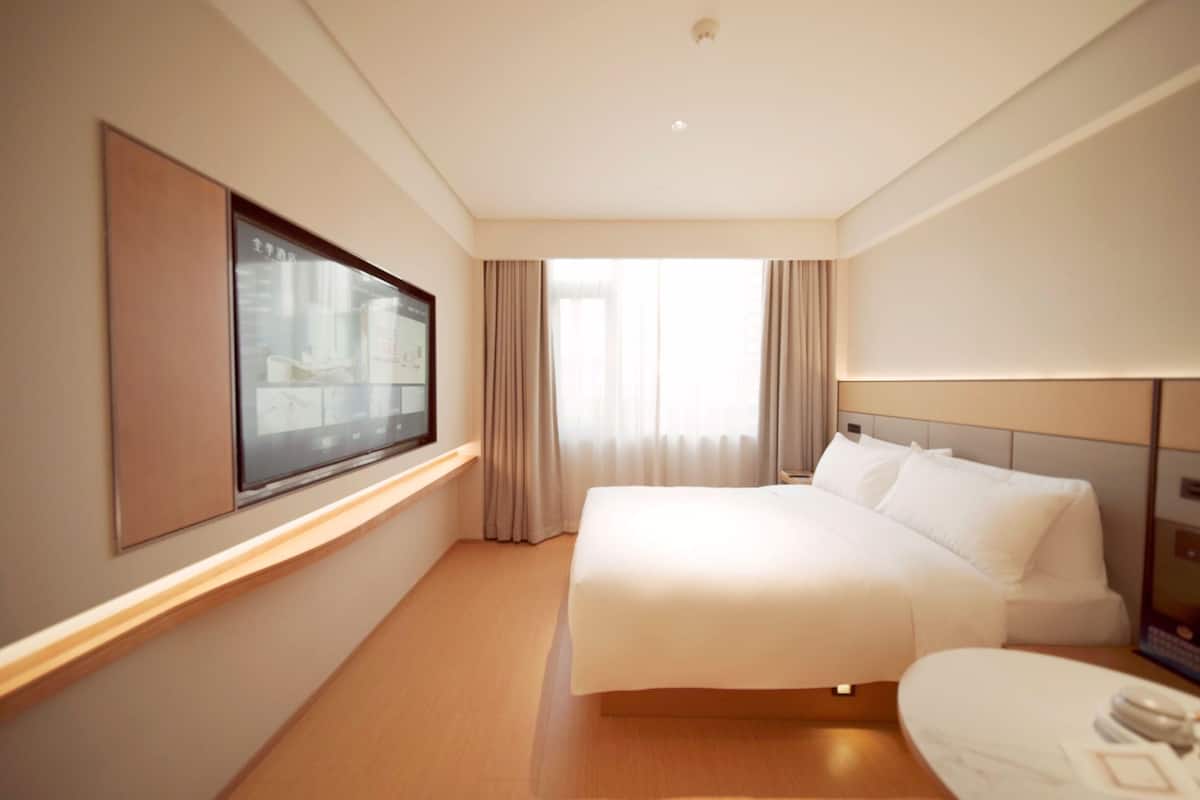 A hotel room with a large bed, a flat-screen TV mounted on the wall, and a window with curtains.