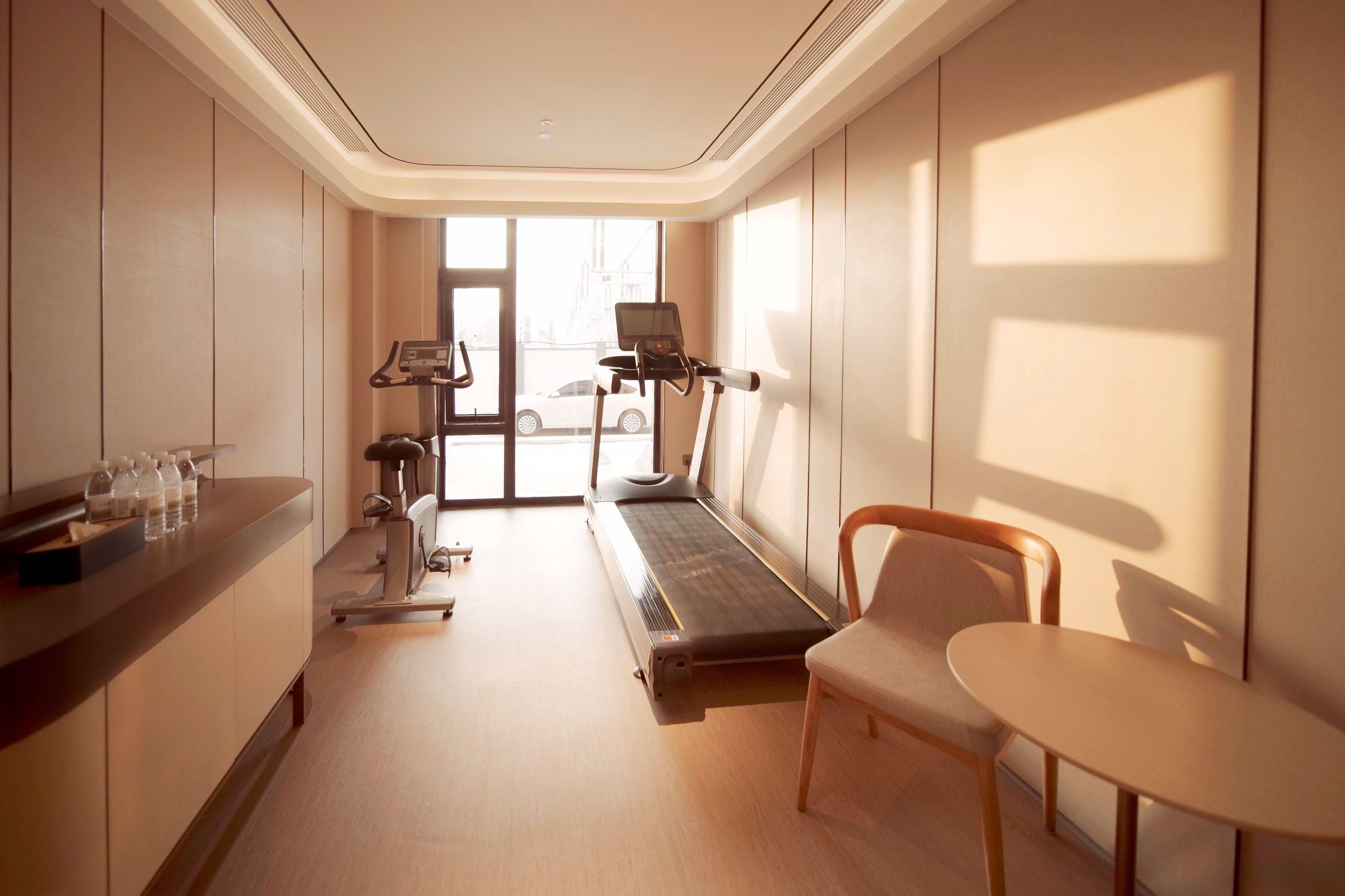 A hotel room with a treadmill, exercise equipment, a desk with water bottles, and a view of a cityscape through the window.