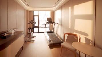A hotel room with a treadmill, exercise equipment, a desk with water bottles, and a view of a cityscape through the window.
