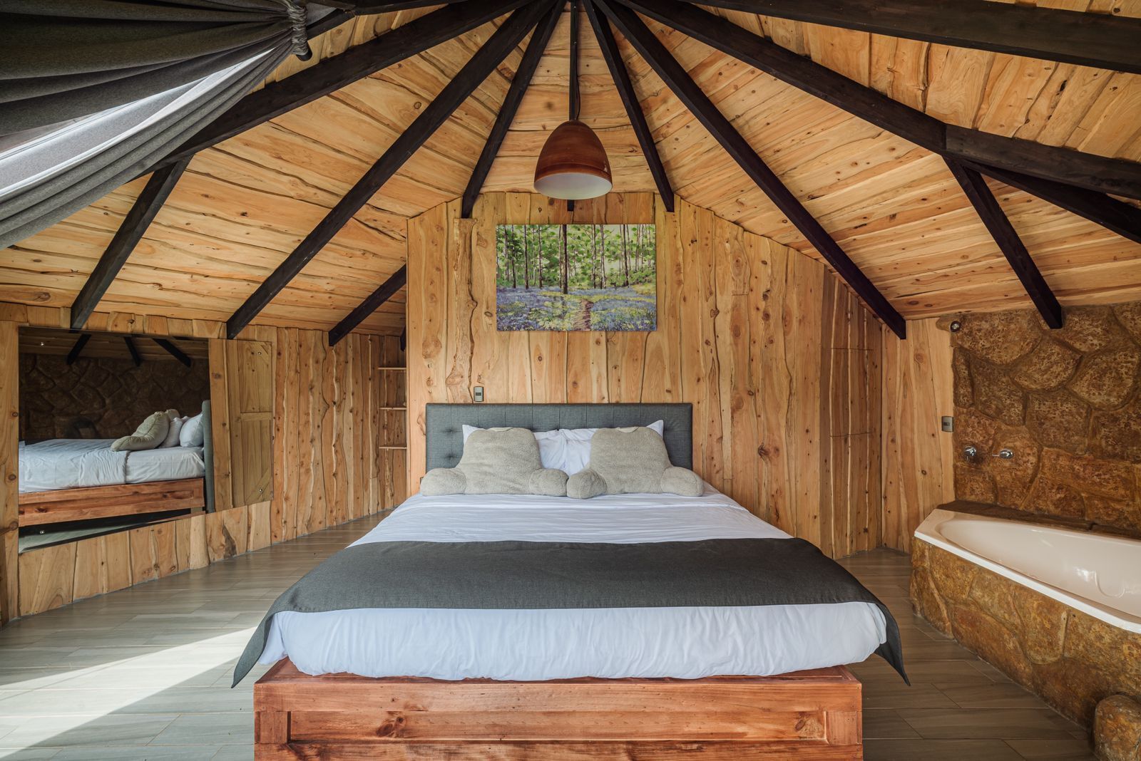 A wooden cabin bedroom with a large bed, a built-in bench, a stone wall, and a window with a painting.