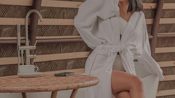 A person sitting on a white chair with a towel, a small table with a cup and saucer, and a metal faucet.