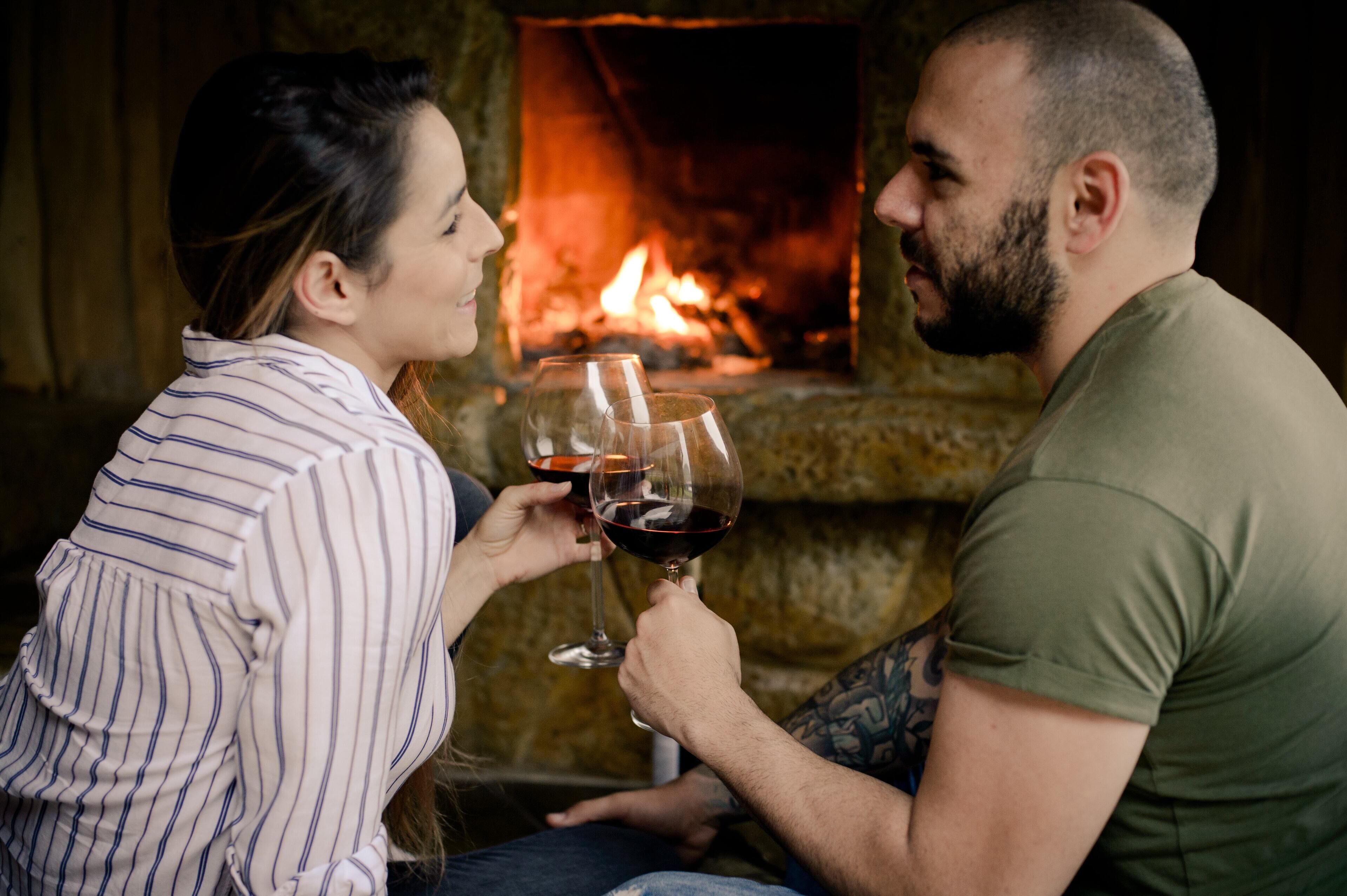 A couple sitting by a fireplace with wine glasses.