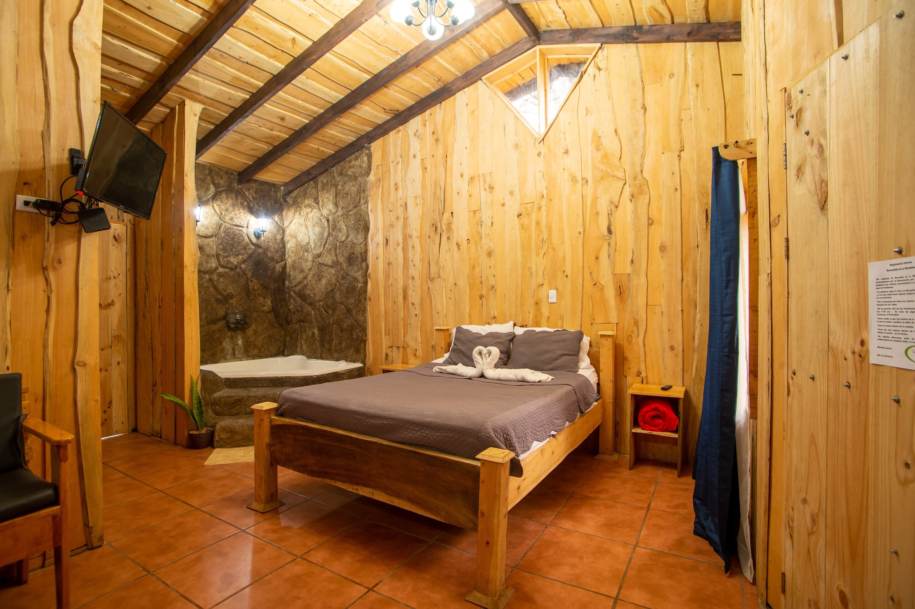 Une pièce de style cabane en bois, avec un lit, un mur en pierre, une cheminée en pierre, un plafond en bois et un téléviseur fixé au mur.