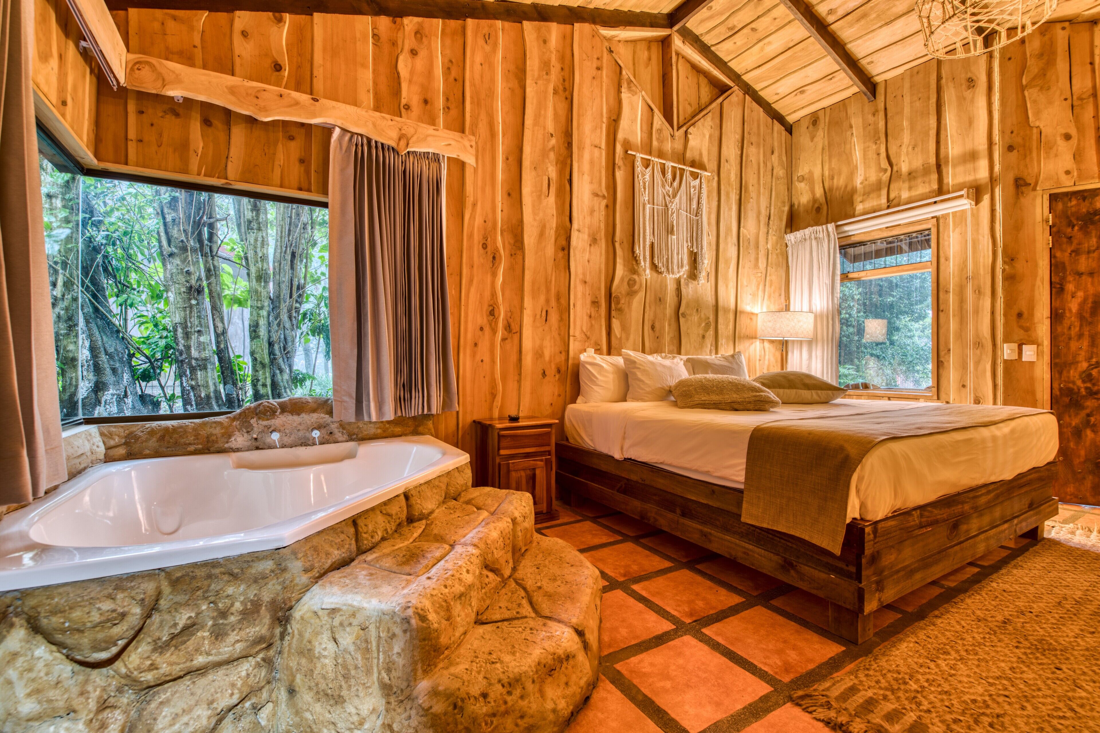 Un dormitorio de cabaña de madera con una bañera de piedra, una cama con sábanas blancas y vistas a árboles por la ventana.