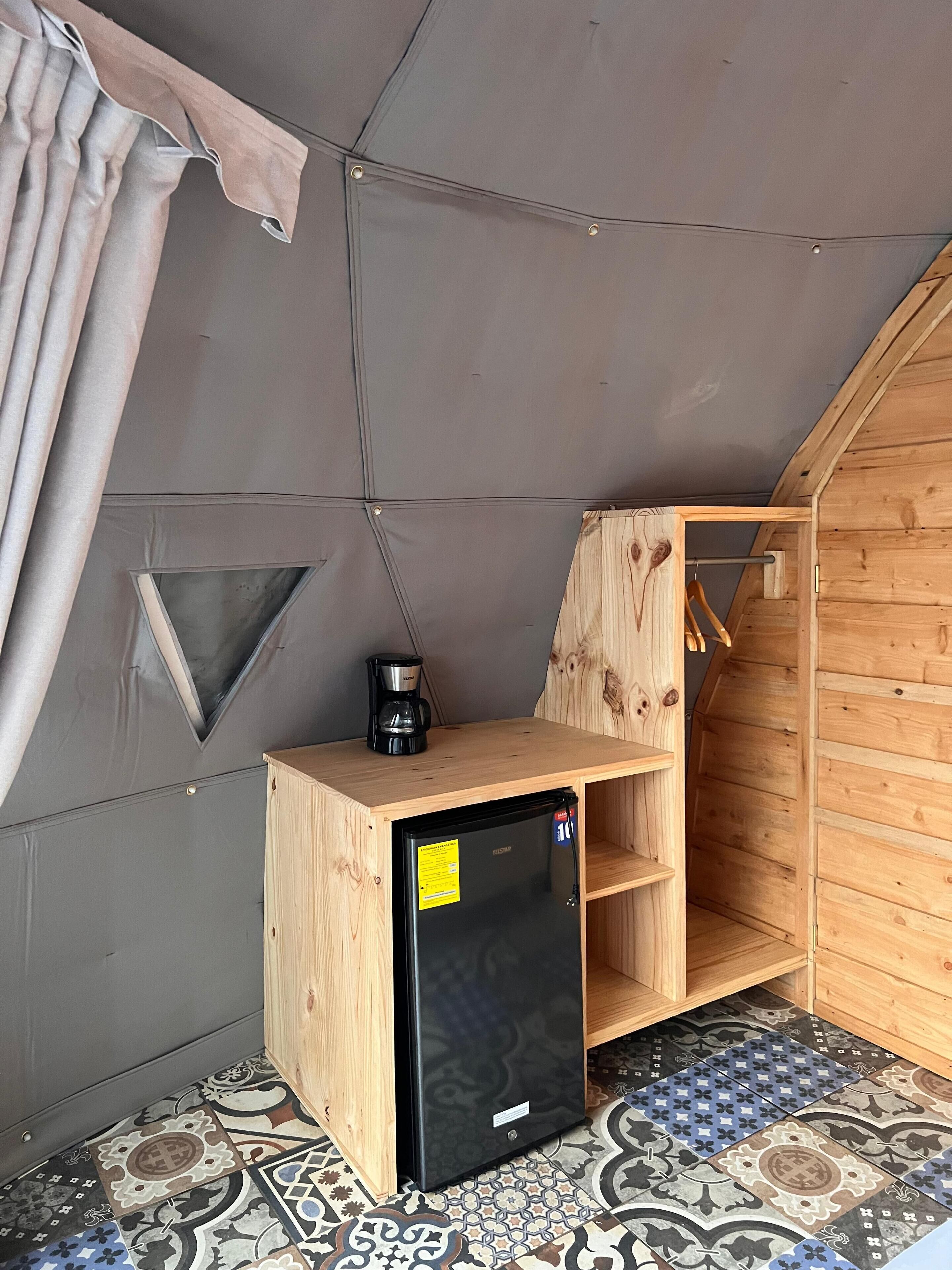 A compact room with a wooden cabinet, a black refrigerator, and a coffee maker on a tiled floor.