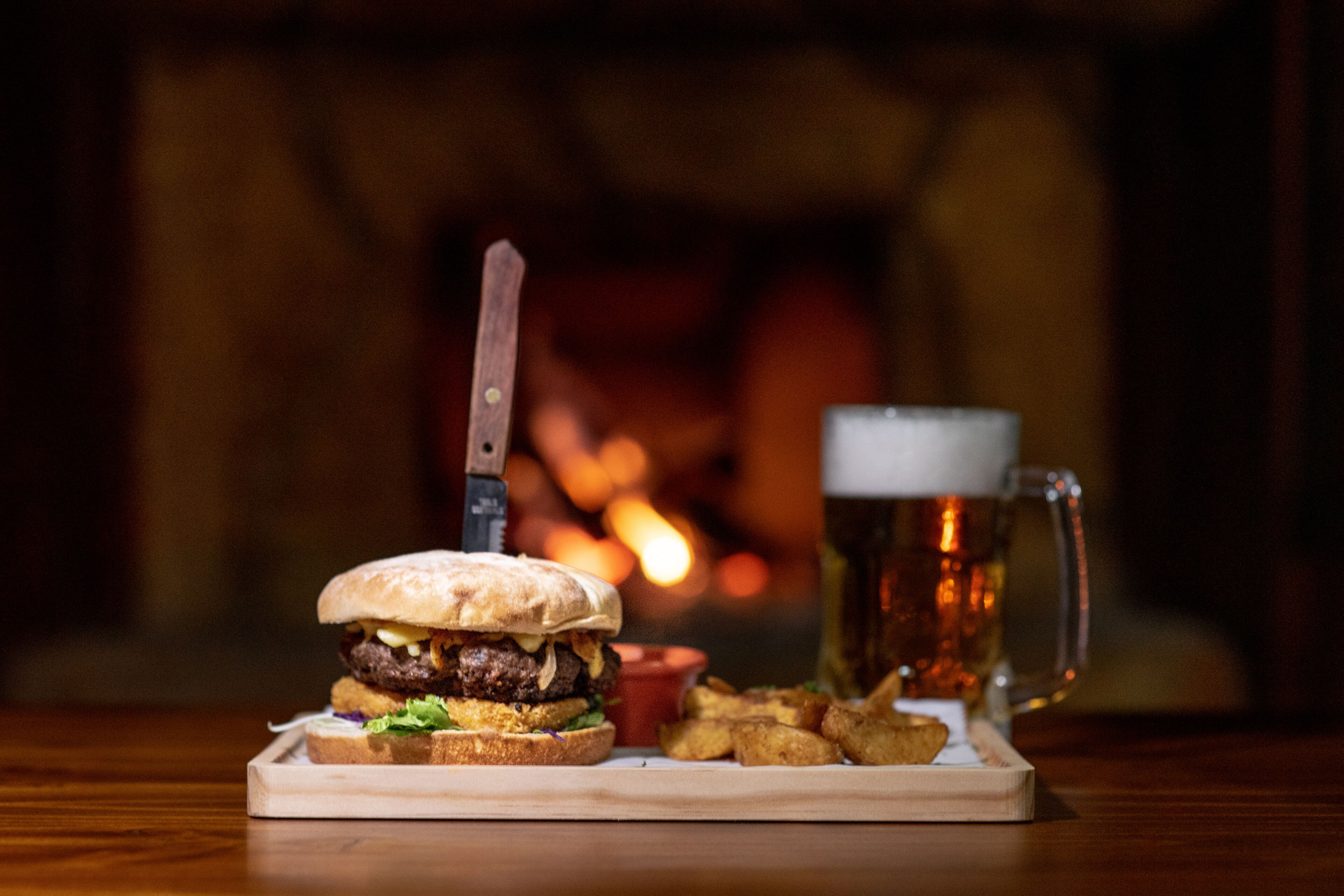 Hamburguesa con un cuchillo sobre una tabla de madera, un vaso de cerveza y una porción de papas fritas.