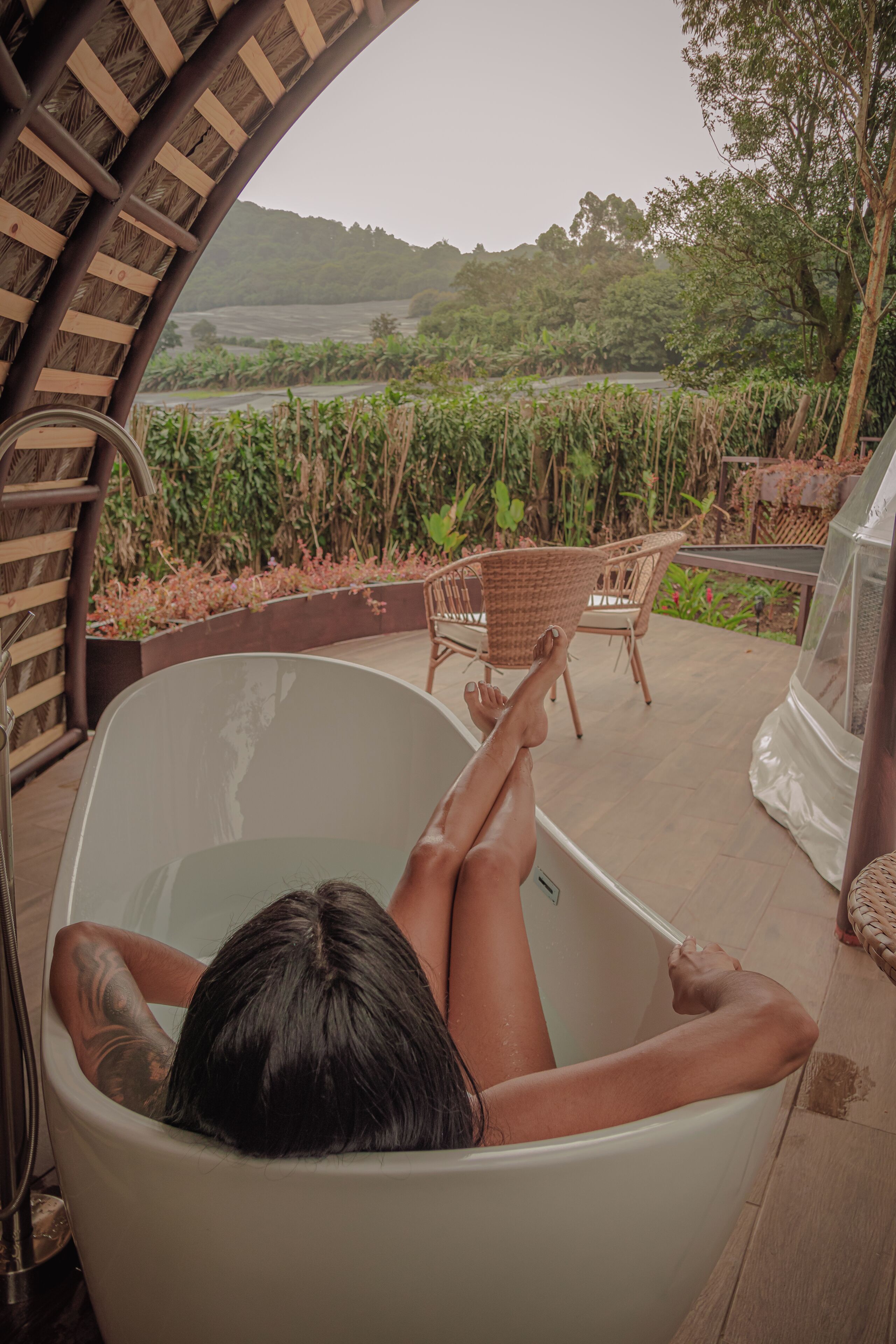 Una persona relajándose en una bañera con vistas a un exuberante jardín y un paisaje distante.