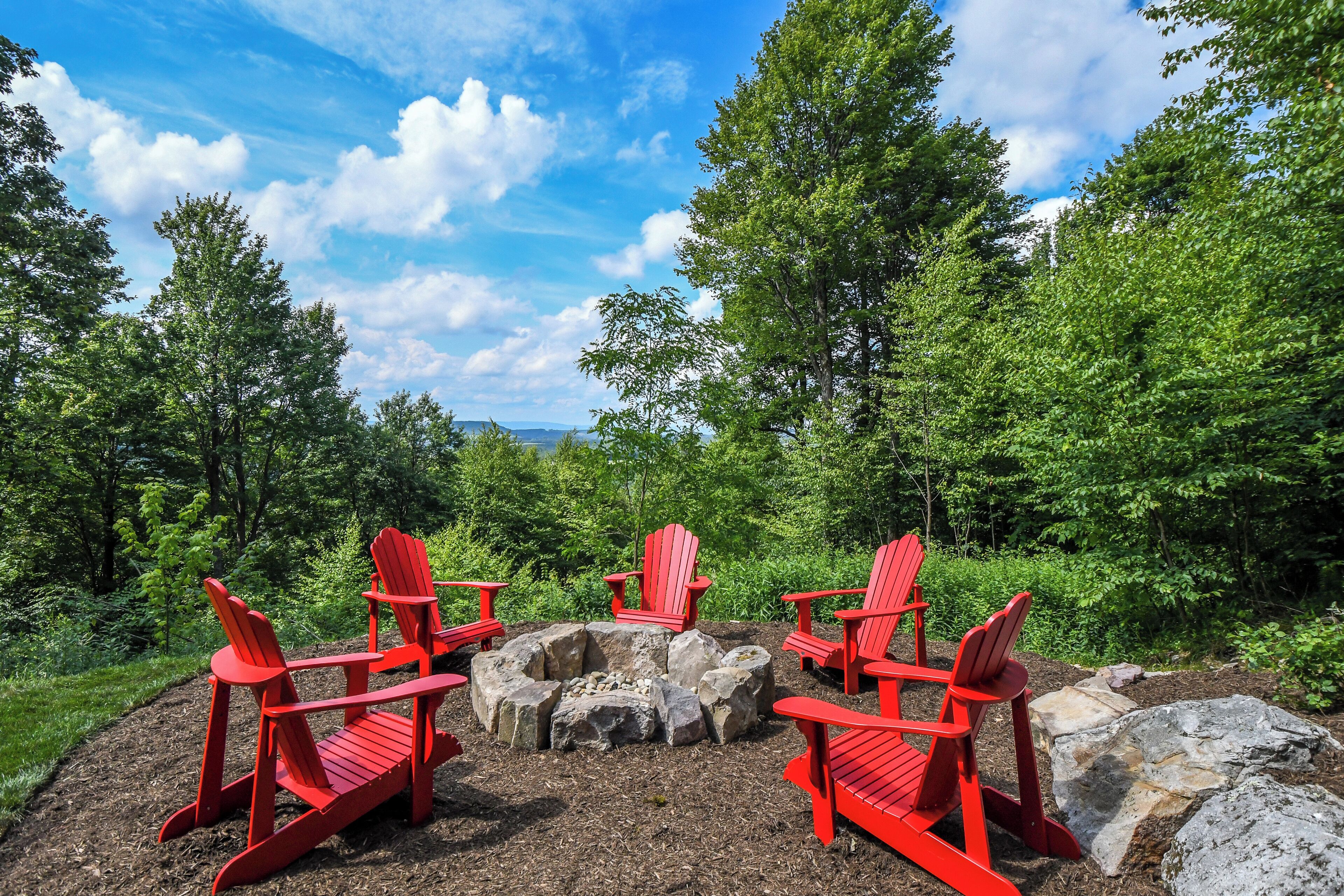 Une zone boisée avec un foyer et des chaises Adirondack rouges.