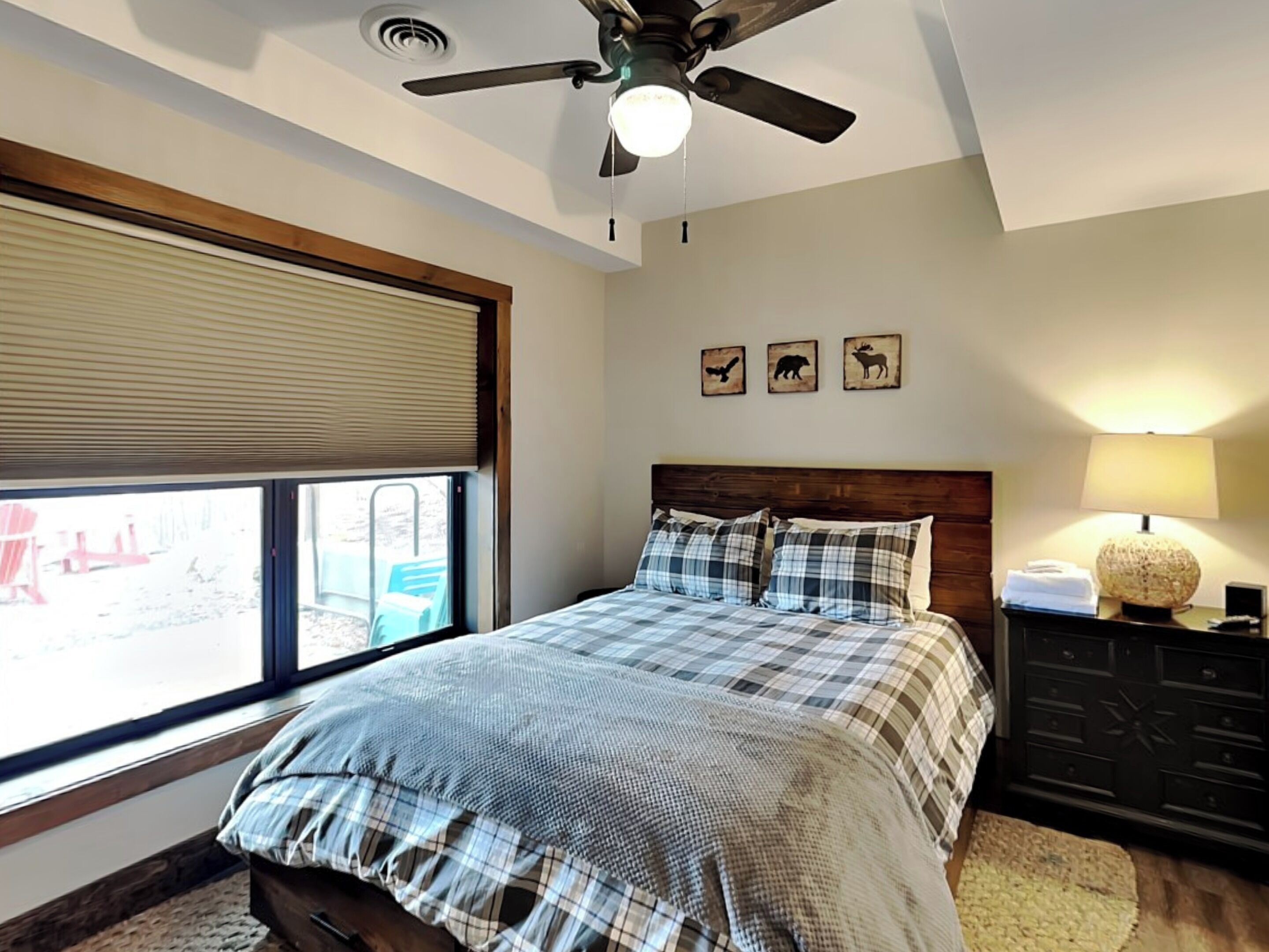 Un dormitorio con cama, ventilador de techo, ventana con persianas, mesita de noche con lámpara y cómoda.