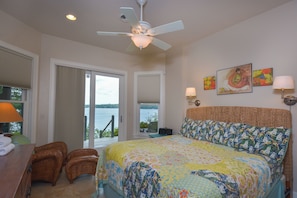 A bedroom with a bed, a ceiling fan, a view of the outdoors, and artwork on the wall.