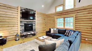 A log cabin interior with a stone fireplace, a large sofa, and a television. - Almost Eden by Taylor Made Deep Creek (McHenry)