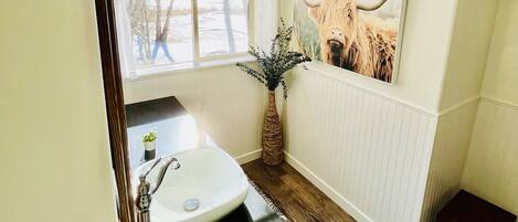 Un baño con un lavamanos blanco, una bañera negra, suelo de madera, una ventana con vistas a árboles y un cuadro de vaca colgado en la pared.