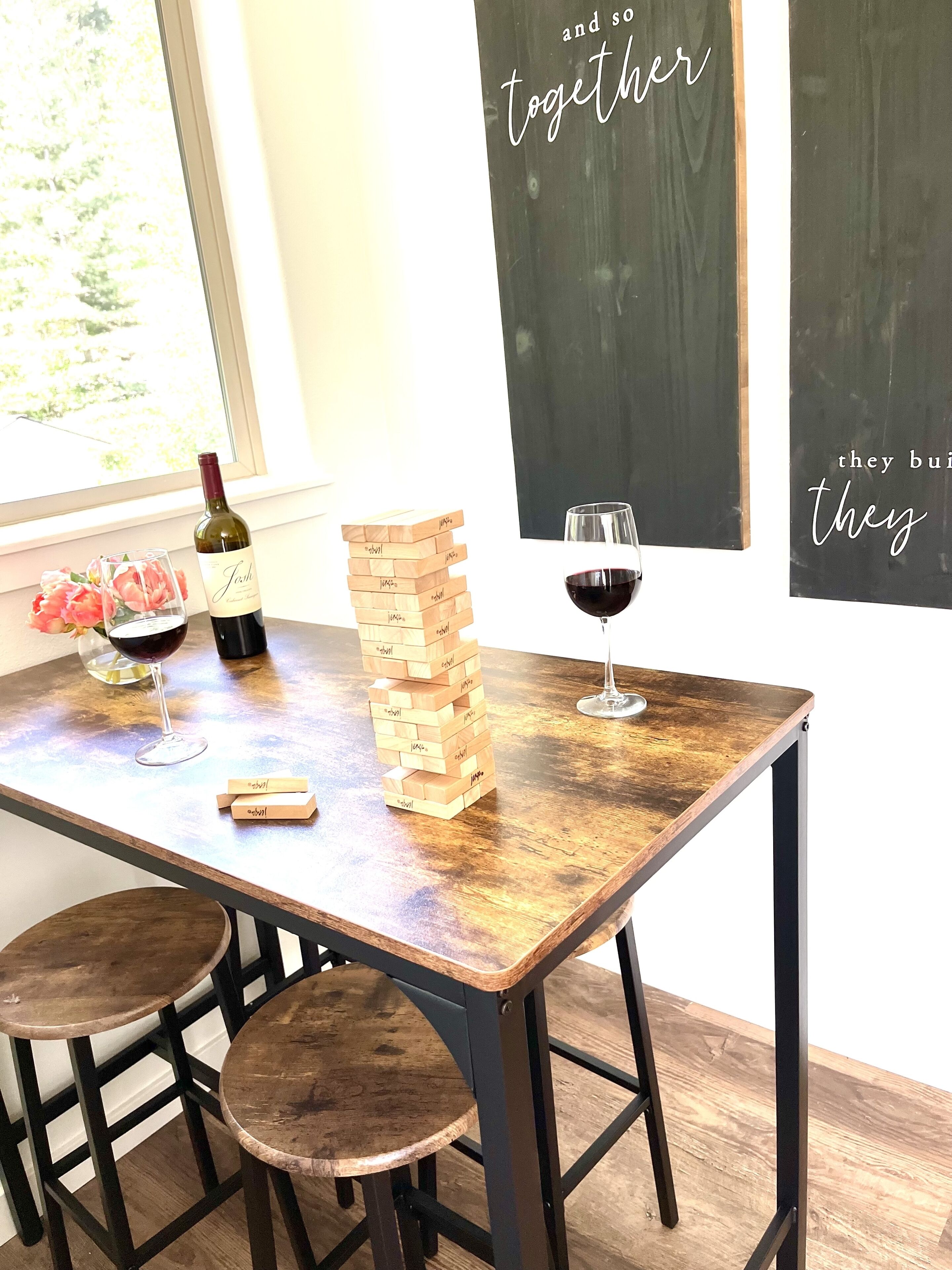 A wooden table with a stack of wooden blocks, a bottle of wine, two glasses of wine, and a single wine glass.